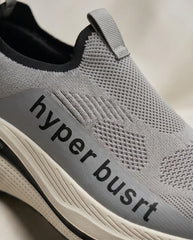 Close-up of a gray slip-on athletic shoe with a black 'hyper busrt' logo on a gray side panel, a black collar, and a cream-colored sole.